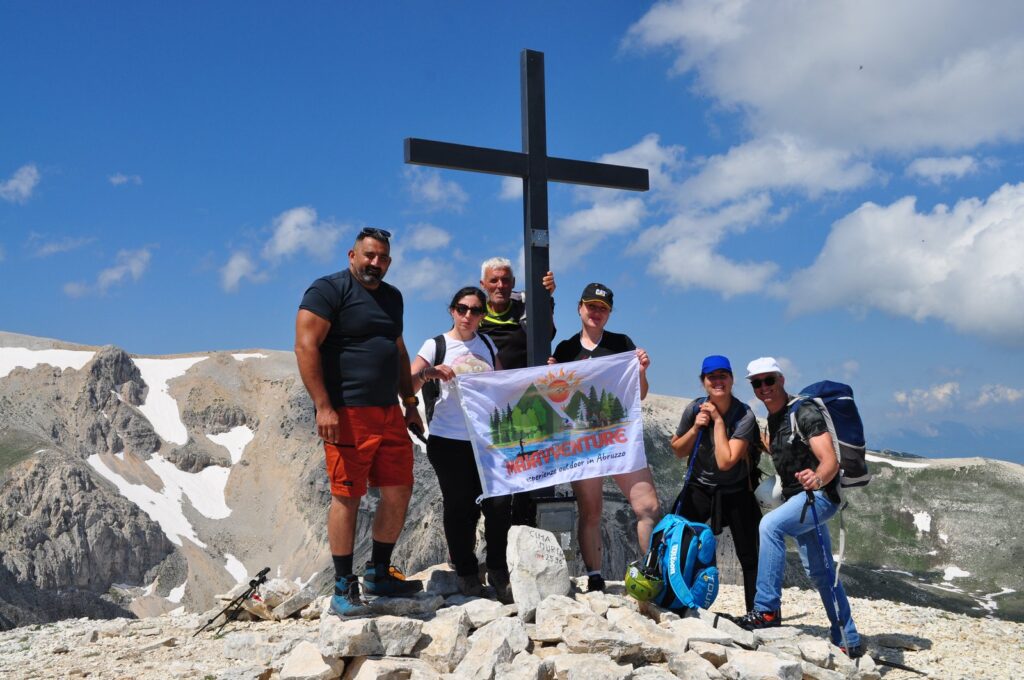 Escursionisti di Majavventure in vetta con la bandiera dell’associazione accanto alla croce